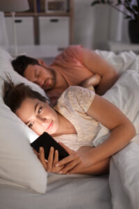 Kobieta trzyma smartfon przed twarzą będąc w łóżku, obok niech leży mężczyzna